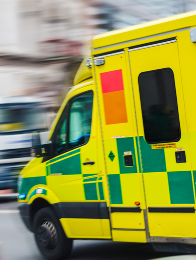 A blurry, side-on shot of a yellow and green UK ambulance speeding down a street. The green checkered pattern is visible on the side of the vehicle, and the motion blur suggests high speed.