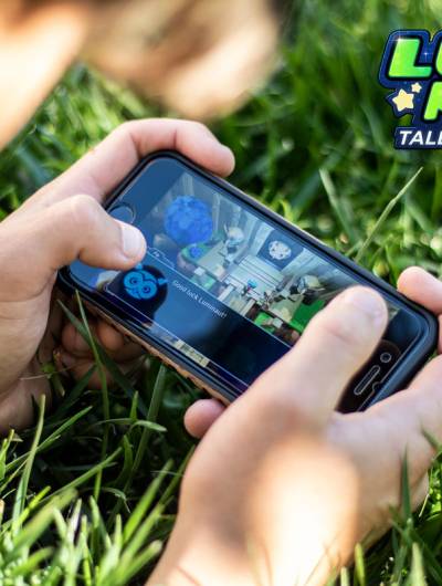 A close-up shot shows a person holding a smartphone horizontally, actively playing a game while sitting in green grass. The game screen displays colourful graphics, and the 